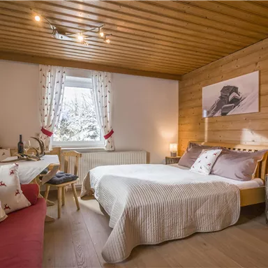 Ein gemütliches Zimmer mit Holzdecke und Holzwänden. Es gibt ein Doppelbett, einen kleinen Sitzbereich und große Fenster, die Tageslicht hereinlassen.