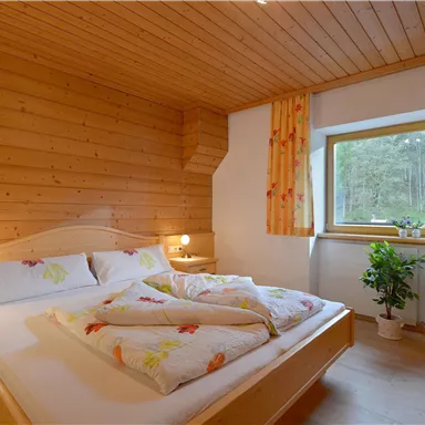 Ein gemütliches Schlafzimmer mit Holzverkleidung und einem großen Bett. Durch das Fenster fällt natürliches Licht und eine Pflanze sorgt für frische Atmosphäre.