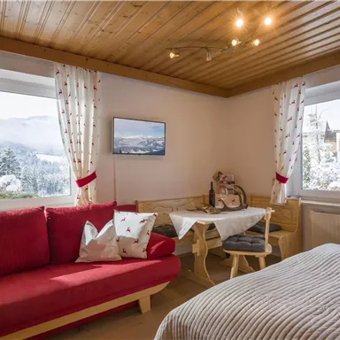 Ein gemütliches Zimmer mit rotem Sofa und einem Bett. Durch das Fenster blickt man auf eine winterliche Landschaft.