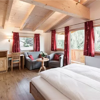 A cozy room with two beds and a wooden ceiling. Large windows with red curtains offer a view of nature.