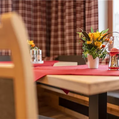 Ein einladender Tisch mit rotem Tischdecken, einem Blumengesteck und einer Glasvase. Im Hintergrund sind gemütliche Fenster und ein kariertes Muster sichtbar.