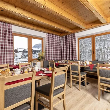 Ein gemütlicher Speisesaal mit Holztischen und Stühlen. Durch die Fenster sieht man die verschneite Landschaft draußen.