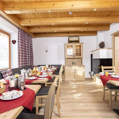 Ein gemütlicher Frühstücksraum mit Holzdecken und großen Fenstern. Tische sind mit frischen Brötchen, Getränken und Geschirr gedeckt.