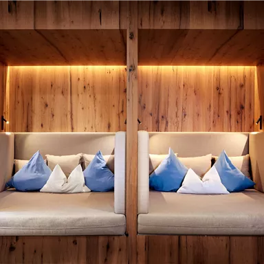 A cozy room with two seating areas made of wood. The seats are decorated with colorful cushions and accentuated by soft lighting.