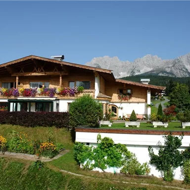 A charming alpine house with flower balconies, surrounded by green meadows. In the background, majestic mountains can be seen.