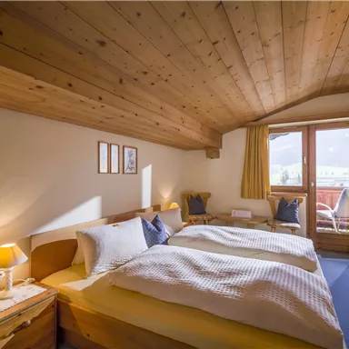 A cozy bedroom with two beds and a wooden ceiling. Large windows provide plenty of daylight and offer a view outside.