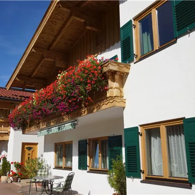 Ein charmantes Haus mit blühenden Balkonpflanzen und grünen Fensterläden. Die Umgebung ist sonnig und einladend.