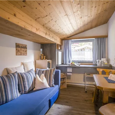 A cozy living space with blue sofa cushions and a wooden shelf. By the window, there is a table with chairs and a small flower vase.
