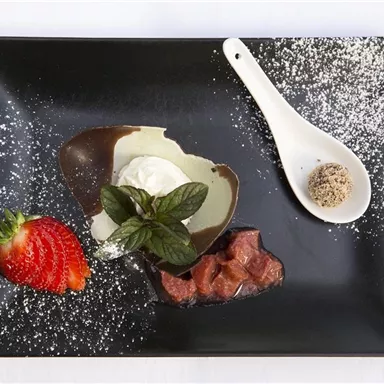 Ein elegantes Dessert auf einem schwarzen Teller, bestehend aus einer Schokoladenkugel, Sahne und frischen Erdbeeren. Dekoriert mit Minze und Puderzucker.