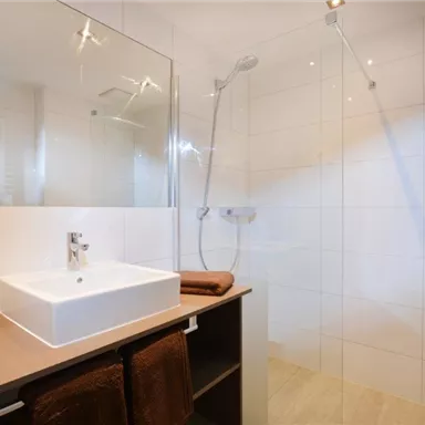 Ein modernes Badezimmer mit einer Dusche und elegischem Waschbecken. Die Wände sind hell gefliest, und es gibt eine stilvolle Ablage.