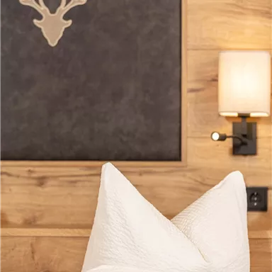 Ein gemütliches Schlafzimmer mit weißen Kissen und einer Holzwand. An der Wand hängt ein Hirschgeweih und eine Wandlampe.