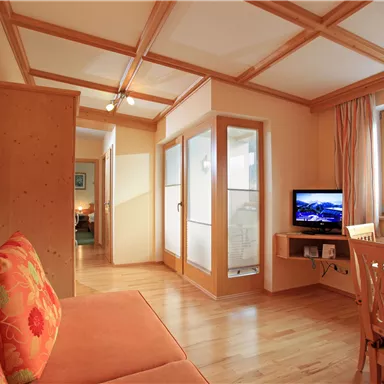 Ein helles Wohnzimmer mit einem gemütlichen Sofa und einem Fernseher. Der Raum ist mit Holzdetails und großen Fenstern ausgestattet, die Tageslicht hereinlassen.