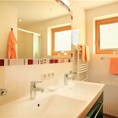 Ein modernes Badezimmer mit einem Doppelwaschtisch und großen Spiegeln. Helle Fliesen und orangefarbene Handtücher verleihen dem Raum eine freundliche Atmosphäre.