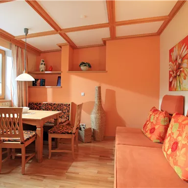 Ein gemütlicher Wohnraum mit einem Esstisch und bunten Stühlen. Die Wände sind in warmen Orangetönen gehalten und mit Blumenbildern dekoriert.