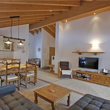 A modern living room with a wooden ceiling and a large dining table. The cozy furnishings consist of a sofa group and a television.
