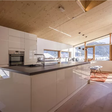 A modern kitchen with white furniture and large windows that let in plenty of daylight. In the background, an impressive mountain landscape can be seen.