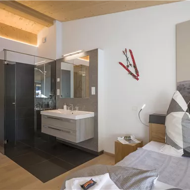 A modern bedroom with wooden accents and an adjoining bathroom. The decor is simple and elegant, featuring a large mirror and a cozy bed.