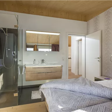 A modern bedroom with a bathroom area. The shower is separated by a glass door and the room feels bright and inviting.