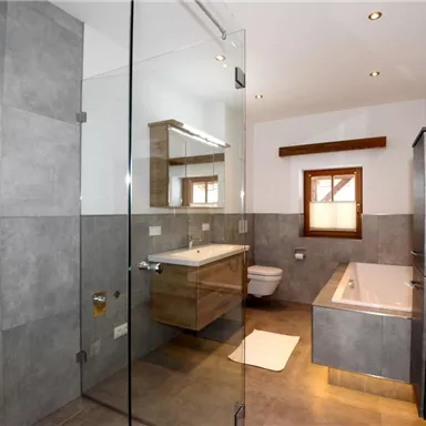 Ein modernes Badezimmer mit grauen Fliesen und einer großen Glasdusche. Es gibt eine Badewanne, einen Waschbereich und einen Holzschrank.