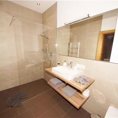 Ein modernes Badezimmer mit einer verglasten Dusche und hellem, neutralem Farbton. Auf dem Holzregal liegen Handtücher und Pflegeprodukte.