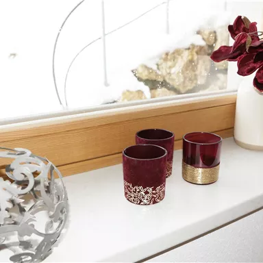 Eine stilvolle Fensterbank mit dekorativen Kerzen und einer Blumenvase. Im Hintergrund sind schneebedeckte Steine sichtbar.