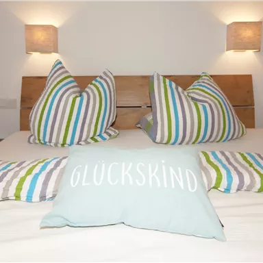 Ein gemütliches Schlafzimmer mit einem Doppelbett und bunten Kissen. Der Schriftzug "GLÜCKSKIND" auf einem Kissen verleiht dem Raum eine freundliche Note.