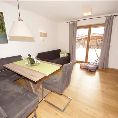 Ein modernes Wohnzimmer mit einem Esstisch und gemütlichen Sofas. Große Fenster lassen viel Tageslicht herein und bieten einen Blick nach draußen.