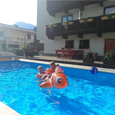 An inviting swimming pool with children swimming on an inflatable toy. In the background, a charming house and the surrounding landscape can be seen.