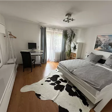 Ein modernes Schlafzimmer mit einem großen Bett und grauen Bettdecken. Es gibt einen Schreibtisch, einen Fernseher und große Fenster, die viel Tageslicht hereinlassen.