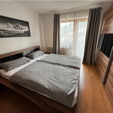 Ein Schlafzimmer mit zwei Einzelbetten und grauer Bettwäsche. Große Fenster lassen viel Tageslicht herein und es gibt Holzregale an der Wand.