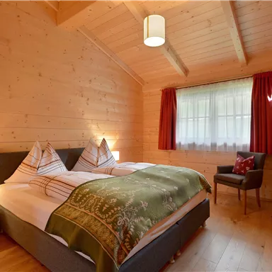 A cozy room with wooden paneling, two single beds, and a green throw. There is a television, a chair, and large windows with red curtains.