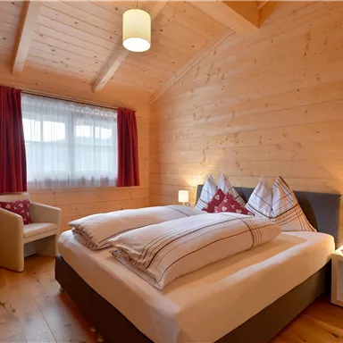 A cozy bedroom with wood paneling and a double bed. There is a television, an armchair, and a window with curtains.