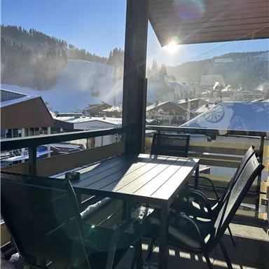 Ein Balkon mit einem Tisch und Stühlen, der einen Blick auf schneebedeckte Berge bietet. Die Sonne scheint klar über die Landschaft.