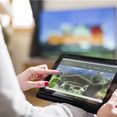 Eine Person hält ein Tablet und schaut auf eine Webseite. Im Hintergrund ist ein Fernseher zu sehen.