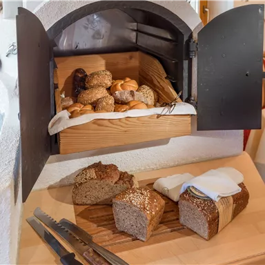 Ein Holzofen mit frisch gebackenem Brot und Brötchen. Auf einem Holzbrett liegt geschnittenes Brot mit einer Serviette.
