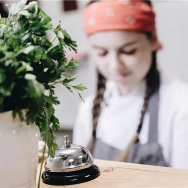 Eine Köchin mit einem roten Kopftuch arbeitet in einer Küche. Im Vordergrund steht ein Blumentopf mit frischer Petersilie.
