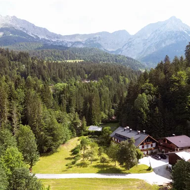 Eine malerische Landschaft mit Bergen im Hintergrund und dichten Wäldern. Im Vordergrund befindet sich ein hübsches Gebäude inmitten von grünen Wiesen.