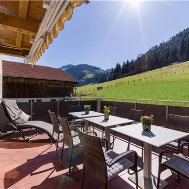 Eine gemütliche Terrasse mit Gartenmöbeln und Tisch. Im Hintergrund erstrecken sich grüne Wiesen und Berglandschaften unter einem klaren Himmel.
