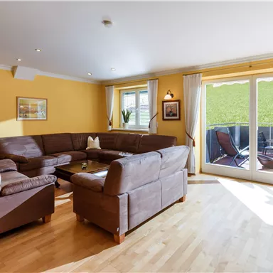 Ein heller Wohnraum mit braunen Ledersofas und einem großen Fenster. Im Hintergrund sind eine Terrasse und Garten sichtbar.