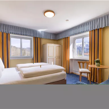 Ein gemütliches Hotelzimmer mit einem Doppelbett, einem Tisch und Stühlen. Große Fenster sorgen für viel Tageslicht und eine angenehme Atmosphäre.