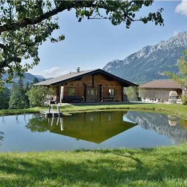 Ein charmantes Holzhaus mit einem klaren Teich im Vordergrund. Die umliegende Landschaft ist grün und von Bergen umgeben.
