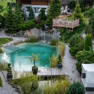 Ein ruhiger Garten mit einem klaren Schwimmteich und viel Grün. Im Hintergrund sind Holzterrassen und Bäume zu sehen.