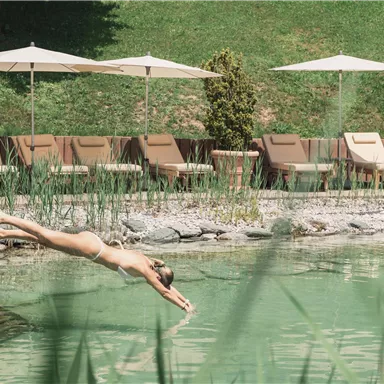 Ein Schwimmer springt in einen klaren Teich. Im Hintergrund sind Liegen und Sonnenschirme zu sehen.