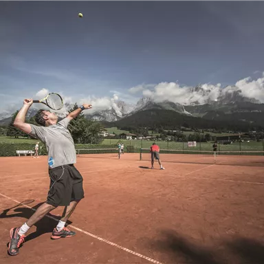 A tennis player hits a ball on a red clay court. In the background, there are mountains and a clear sky.