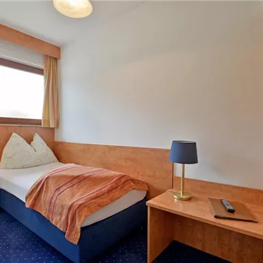 A simple room with a single bed and wooden decor. There is a table with a lamp and a window that lets in daylight.