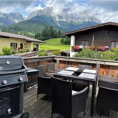 Eine Terrasse mit einem Grill und einem Tisch, umgeben von bequemen Stühlen. Im Hintergrund sind beeindruckende Berge unter einem teilweise bewölkten Himmel zu sehen.