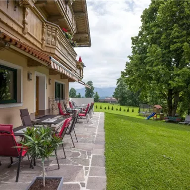 Eine Terrasse mit roten Stühlen und Tischen neben einem gepflegten Garten. Im Hintergrund sind Bäume und eine sanfte Landschaft zu sehen.