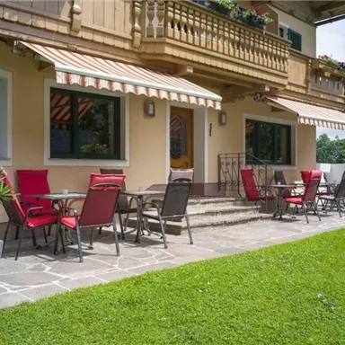 Eine schöne Terrasse mit roten Stühlen und Tischen vor einem Gebäude. Die Umgebung ist grün und einladend.