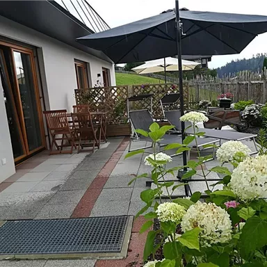Eine gemütliche Terrasse mit Gartenmöbeln und einem Sonnenschirm. Schön dekoriert mit blühenden Pflanzen und Blick auf eine grüne Landschaft.