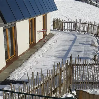 Ein modernes Haus mit einem schneebedeckten Garten. Der Bereich ist von einem Holzzaun umgeben und es gibt eine Terrasse vor den Fenstern.